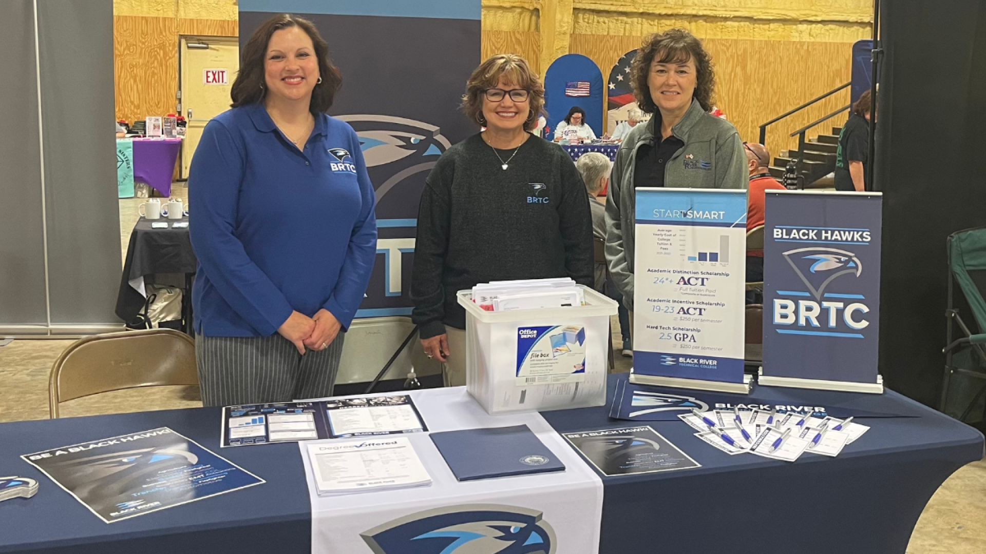 Three women in BRTC apparel stand smiling behind a recruitment table decorated with Black Hawks banners, scholarship information, and college brochures at an event.
